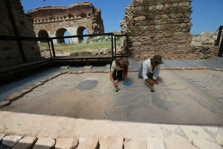 Two conservators repair a mosaic floor in a ruined stone courtyard under a blue sky.