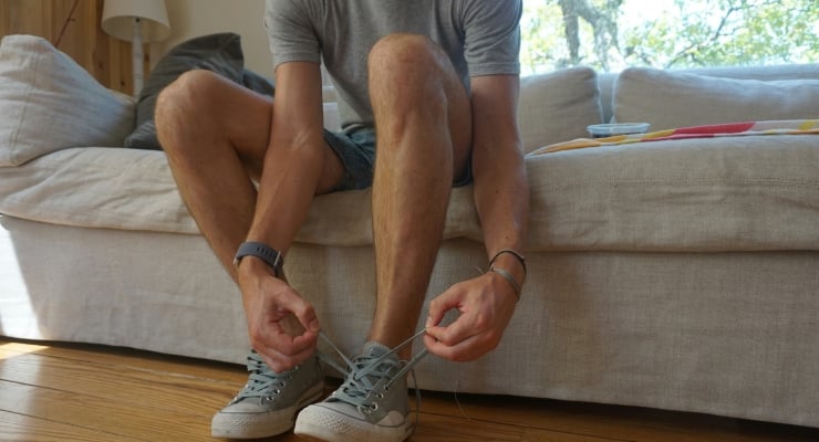 Person sitting on a couch tying the laces of gray sneakers on their feet.