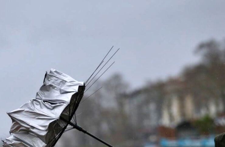 Person in a dark jacket grips a crumpled silver umbrella pushed sideways by strong wind at a windy coastal scene.