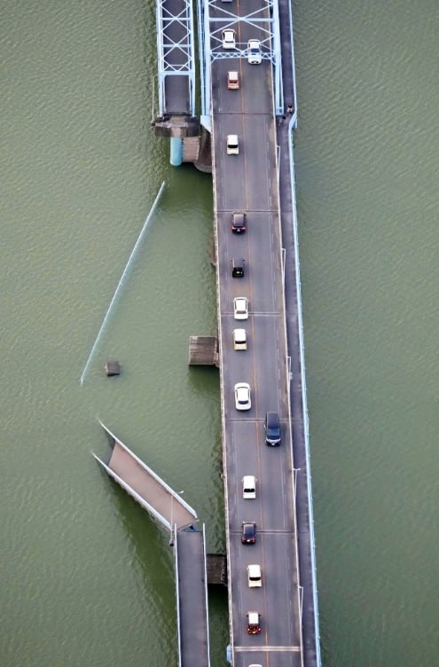 Bird's-eye view of a long bridge over a greenish river with several cars driving along; steel truss supports visible on the left and a lower ramp at the bottom left.