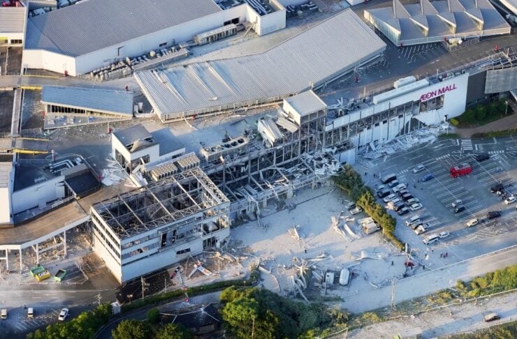 Aerial view of a large shopping mall with extensive structural collapse, rubble scattered in the parking lot and around the damaged facade.