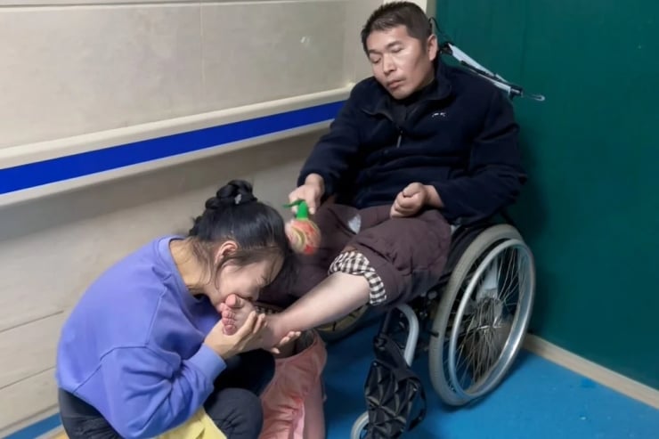 Man in a wheelchair in a hallway being comforted by a woman kneeling beside him, holding his foot.