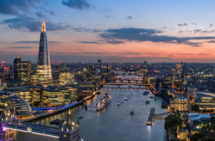 Aerial view of London's skyline at dusk, with Tower Bridge illuminated over the River Thames and modern buildings in the background.