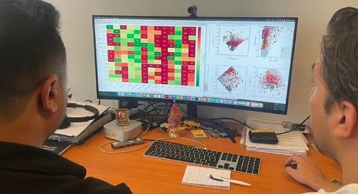 Two people study a data dashboard on a large monitor showing a color-coded heatmap and scatter plots on a desk setup.