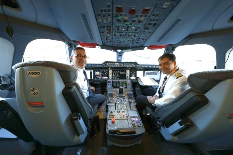 Two pilots sit in the cockpit of an Airbus A350, smiling at the camera with the flight deck instruments ahead.