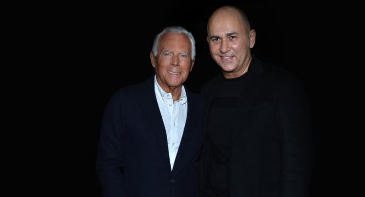 Two smiling men stand side by side against a dark background; one older with white hair wearing a navy blazer and white shirt, the other younger bald man in black.