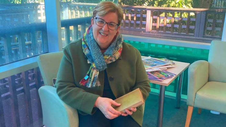 Smiling woman in a green coat and colorful scarf sits in a bright room, holding a small book.