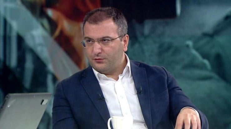 Man in a navy suit and white shirt sits on a TV studio set, wearing glasses and a lapel mic, looking to his left.