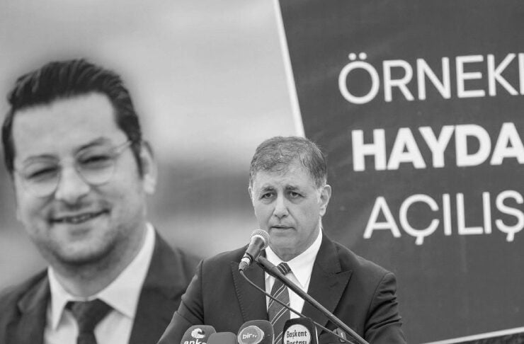 Man in a suit speaks at a podium with multiple microphones during a press conference, with a large banner behind him.