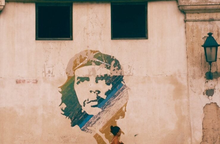 Woman in a red dress walks along a weathered beige wall with a large mural of a man's face beside a street sidewalk.