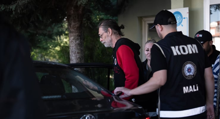 Group of people inspect a black car at a roadside area; a man in a red-sleeved hoodie leans toward the car while others, including an officer with a 'KOM MALİ' vest, stand nearby near a doorway.