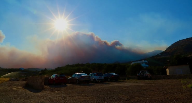 Sun over a smoky hillside with a blue sky, smoke plumes drifting across the landscape, and several parked cars on a dirt lot in the foreground.