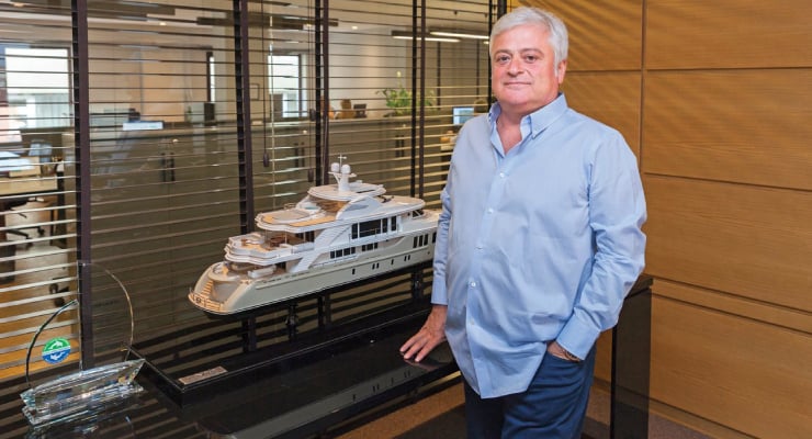Man in a light blue shirt stands beside a scale model yacht on a glass display in a modern office.