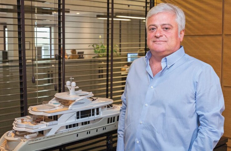 Older man in a light blue shirt stands beside a white luxury yacht model on display in a modern office.