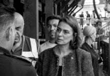 Woman in a textured blazer talks with a uniformed officer among a small crowd in a busy outdoor facility (black and white).