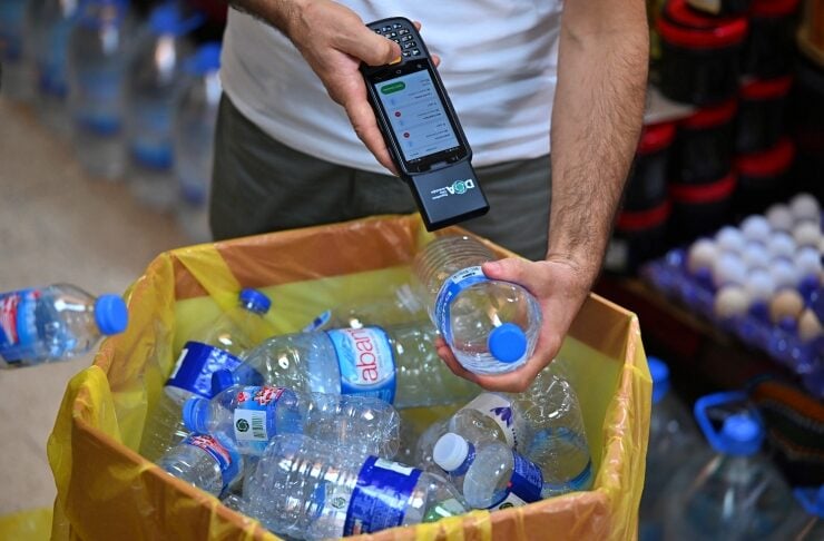 A person scans a plastic bottle with a handheld barcode scanner over a yellow recycling bin filled with plastic bottles.