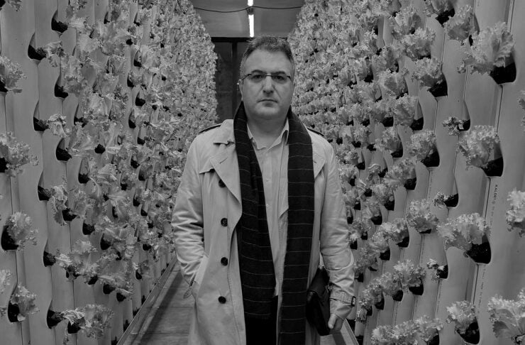 Man in a trench coat and scarf walks down a narrow aisle between vertical rows of leafy crops in a greenhouse.