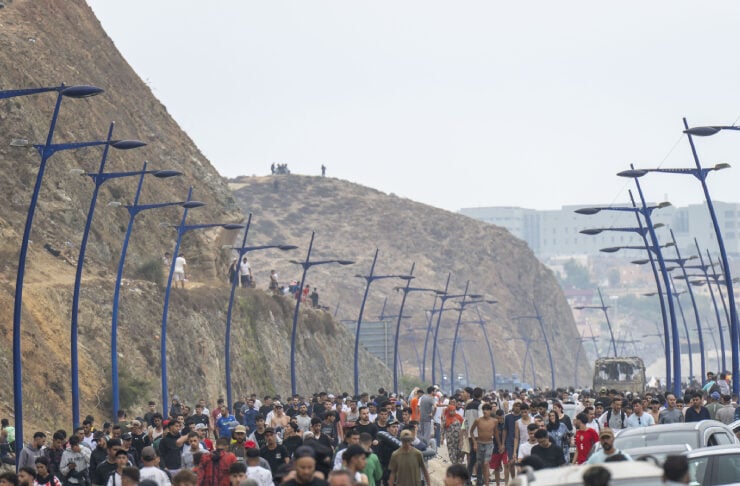 Crowd of pedestrians walking along a beachfront promenade with blue curved street lamps and parked cars on the side.