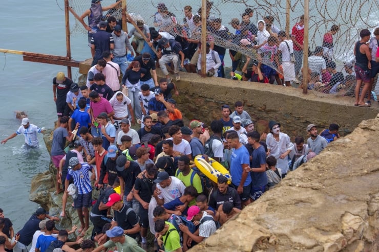 Large crowd of people gathered on a rocky shoreline beside a barbed-wire fence, some wearing life jackets, waiting near the water.