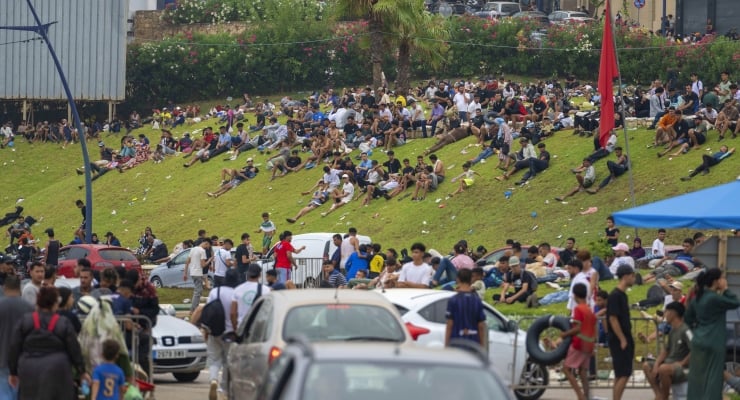 Large crowd sitting on a grassy hill at an outdoor event, with cars parked at the bottom and people enjoying the day.