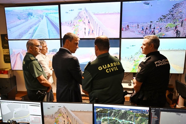 Five officers and security personnel stand in a control room, watching multiple CCTV screens showing coastal barriers, beaches, and water.