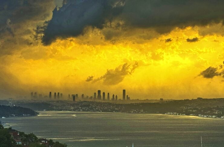 Golden sunset over a distant city skyline across a bay, with stormy yellow clouds and a hillside foreground of houses and trees, including a mosque minaret.