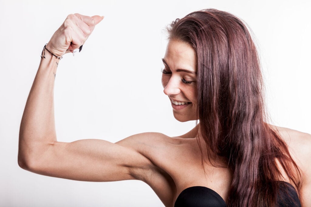 Side-profile of a smiling woman with burgundy hair, flexing her left bicep against a white backdrop with a black strapless top visible at the chest.