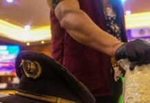 Captain's hat on a table with a white cloth, while a gloved hand holds a clear bag containing white crystalline material in the foreground; blurred colorful lights in the background.
