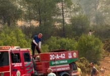 Firefighting crew unloads a red water truck in a smoke-filled forest during a wildfire, with responders nearby and trees in the background.