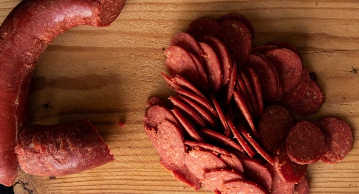 Sliced salami arranged in a neat pile on a wooden cutting board with a curved whole salami on the left.