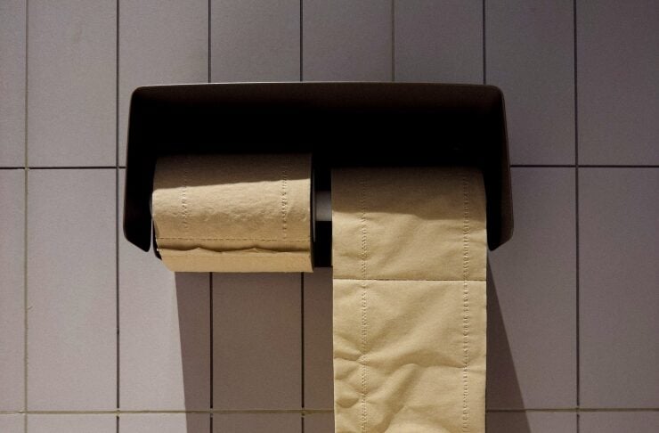 Wall-mounted toilet paper dispenser with a long beige sheet hanging down against a tiled wall.