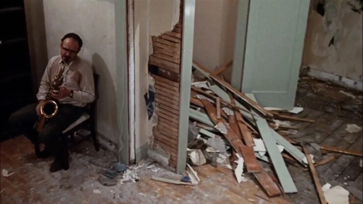 Man playing a saxophone in a dilapidated room with collapsed wall and scattered debris beneath peeling plaster home interior