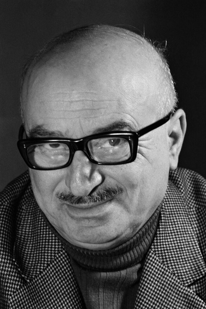 Close-up black-and-white portrait of an older man with thick black glasses, mustache, and a houndstooth jacket looking slightly downward to the left.
