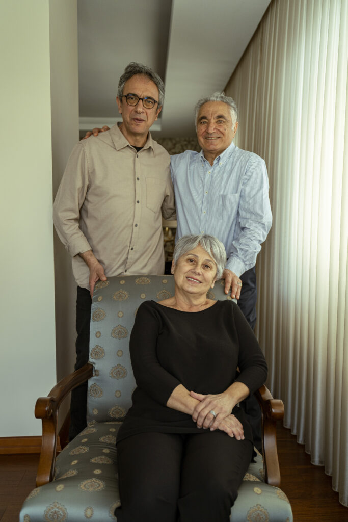 Three older adults pose together in a home setting: a man with glasses in a beige shirt and a man in a blue striped shirt stand behind a smiling seated woman with gray hair on a patterned chair.