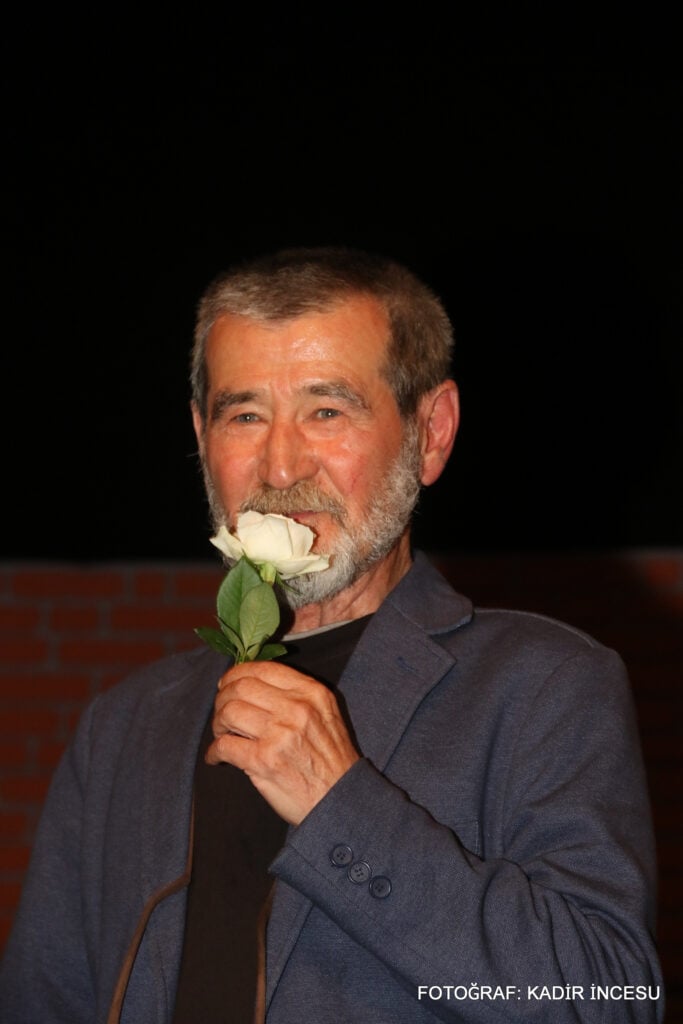 Older man with gray beard holding a white rose near his mouth, wearing a blue blazer, against a dark night background.