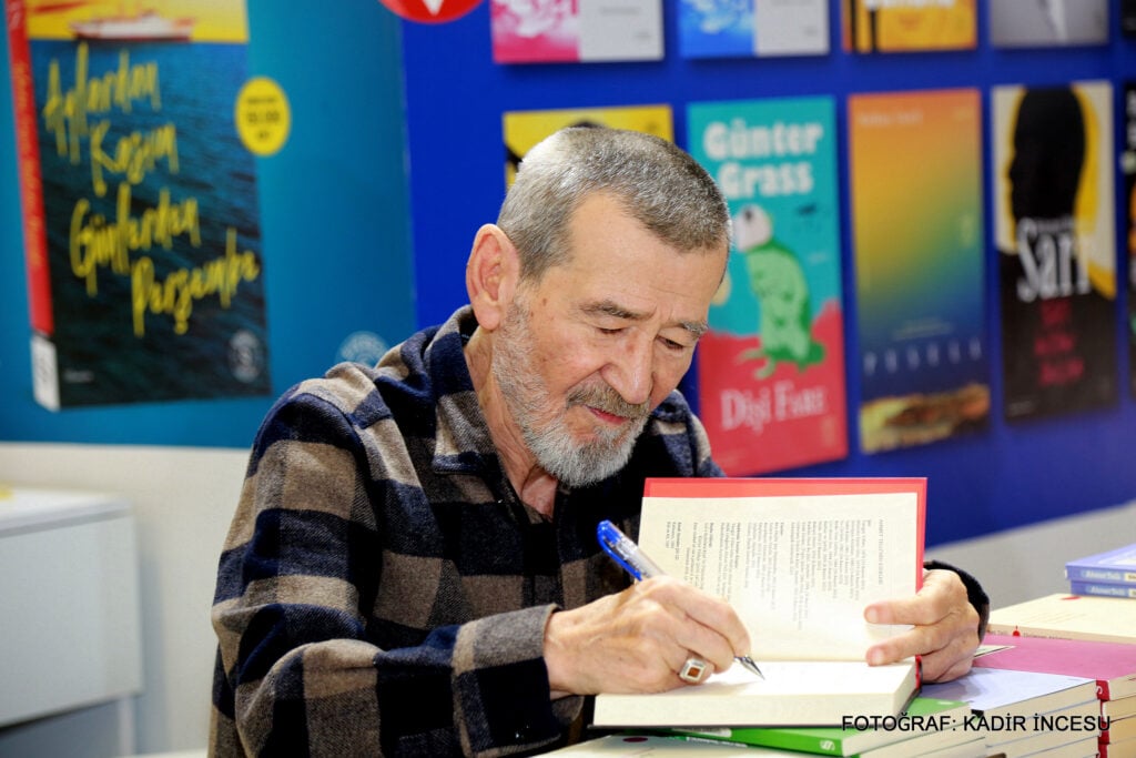 Older man with gray hair signs a book at a table, colorful posters behind him on a blue wall.