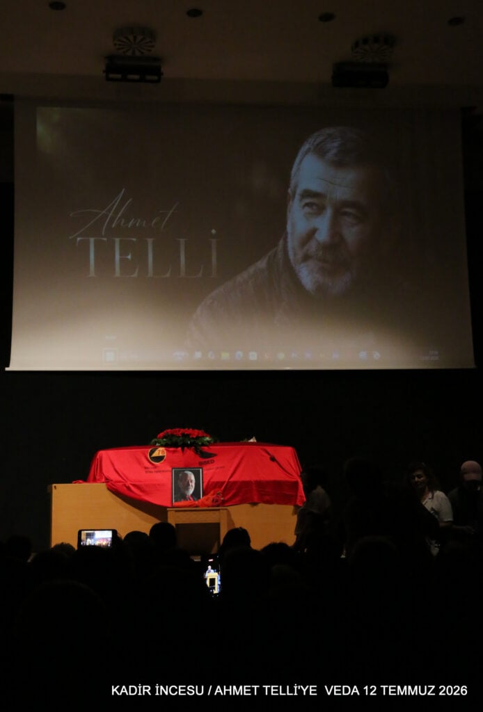 Projection of a man’s portrait on a large screen above a red-draped casket with a small framed photo on the front, during a memorial event.