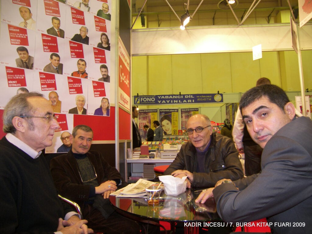 Group of men seated around a glass table at a book fair booth, posters featuring headshots on the wall behind them. Caption text visible: KADIR INCESU / 7. Bursa Kitap Fuarı 2009.