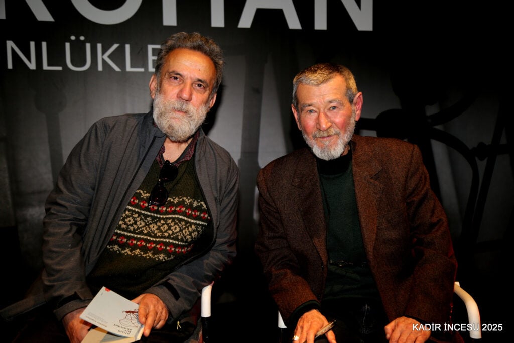 Two older men with gray beards sitting side by side at an event, looking at the camera; man on left wears a patterned sweater and dark jacket, man on right a brown blazer.