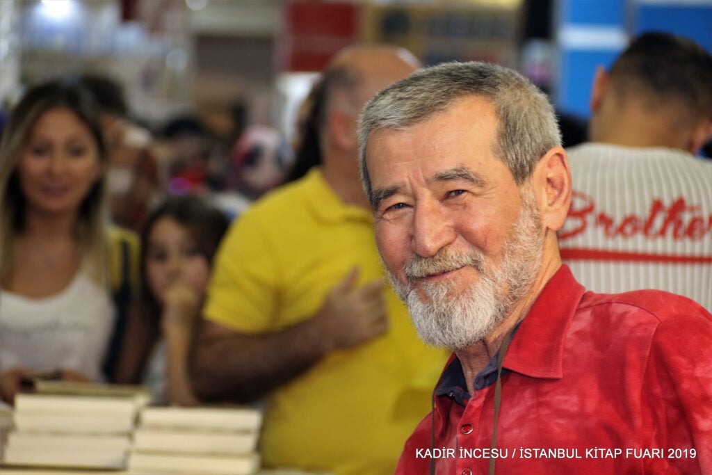 Close-up of a smiling gray-bearded man in a red shirt at a crowded book fair, with stacks of books in the foreground.