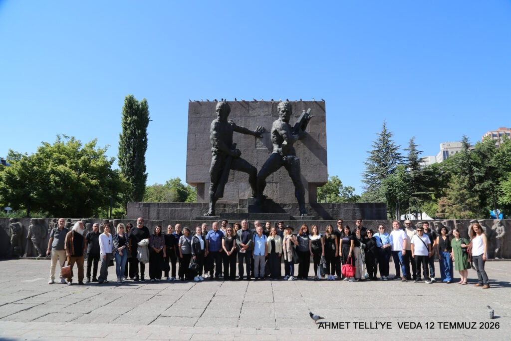 Group of people posing in front of a large dark monument featuring two sculpted figures, outdoors on a sunny day with trees around.