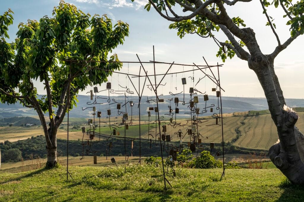 Outdoor metal sculpture made of hanging wire shapes framed by trees on a hillside overlooking fields and hills.