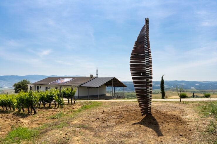 Tall vertical wooden sculpture made of stacked slats in a vineyard, with a single-story building and open countryside in the background.