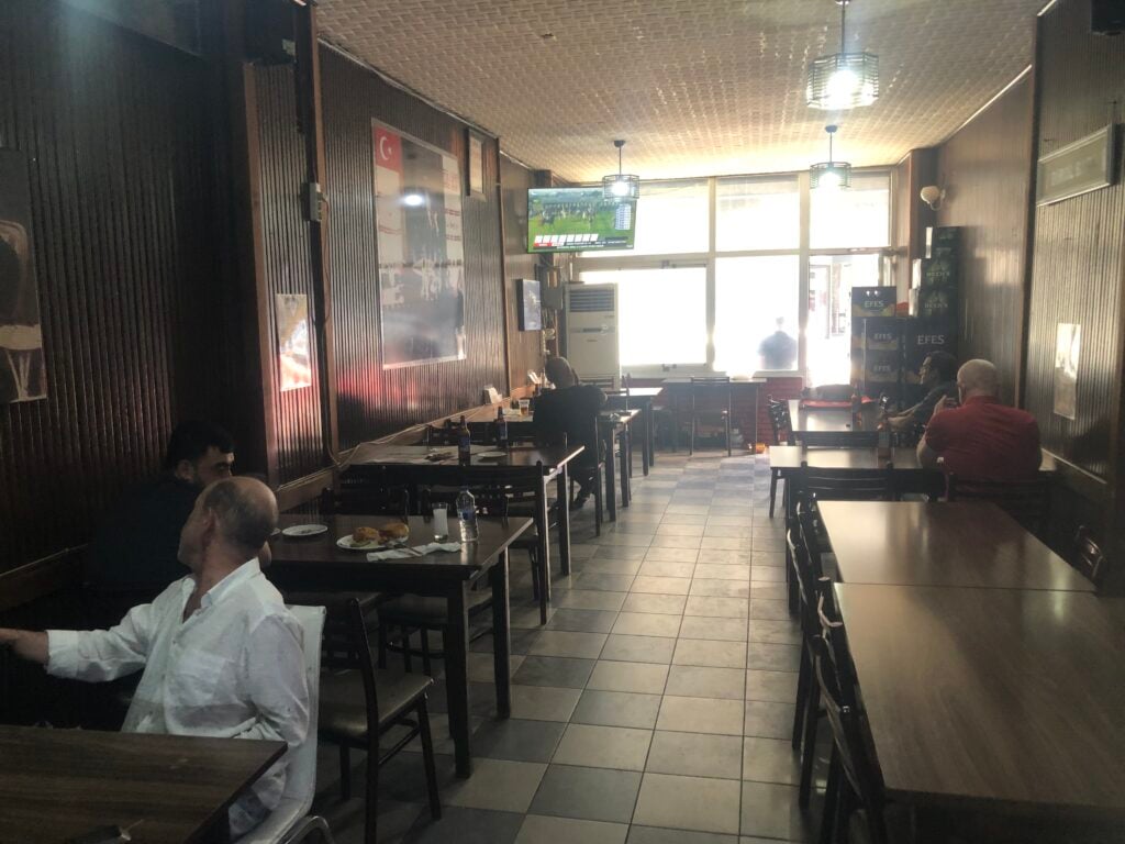 Cafe interior with dark wood walls, tables, and a wall TV showing a sports game. Window light at the back adds brightness.