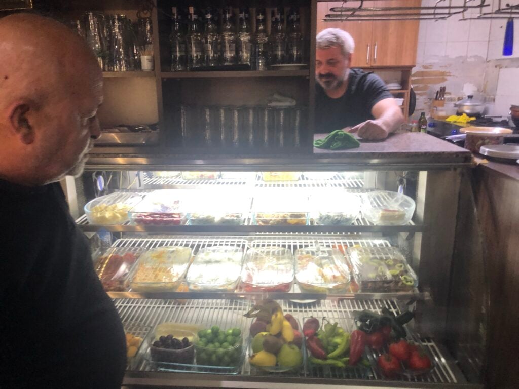 Two men in a deli or kitchen behind a glass counter; fruit cups visible in the display case and shelves of glassware in the background.