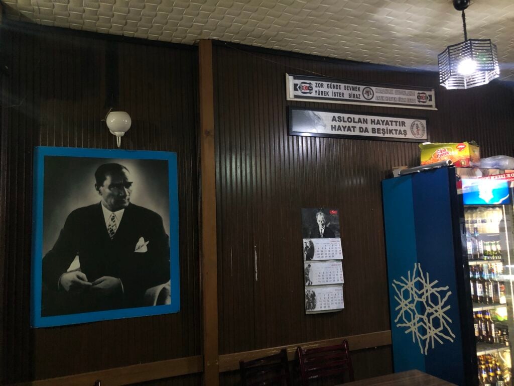 Indoor wall with a large black-and-white portrait in a blue frame, calendar strips, and Turkish banners, plus a vending fridge on the right.