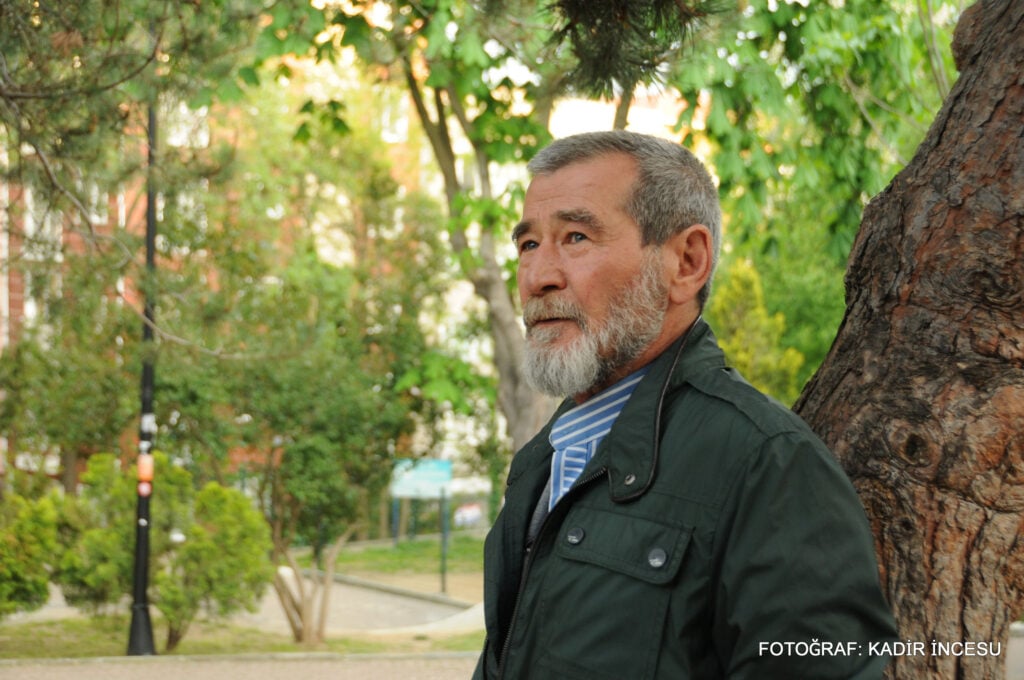 Portrait of an older man with a gray beard standing beside a tree in a sunny park.