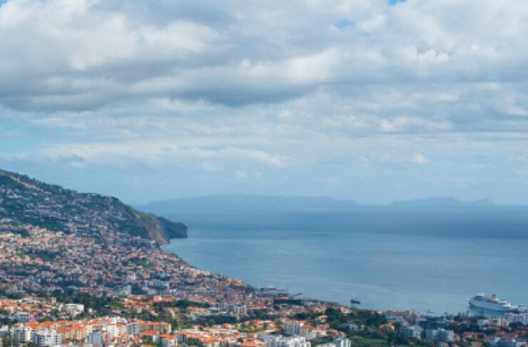 Coastal hillside cityscape with red-roofed buildings cascading toward the blue sea and a distant horizon under a cloudy sky.