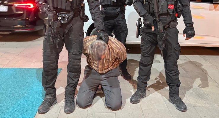 Man kneels with hands behind back while armed officers surround him during arrest on sidewalk at night.