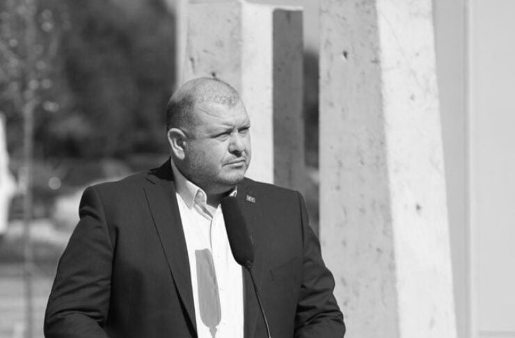 Man in a dark suit giving a speech at an outdoor podium with a microphone and a municipal seal on the lectern (black and white).